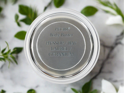 A jar of PëURR® Body Butter showing the product name and the words 'Hyssop / Bay Laurel / Geranium'. The background consists of a marble surface with some leaves scattered around.