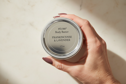 PËURR® Frankincense & Lavender Body Butter held by an older woman with long berry-plum nails against warm marble