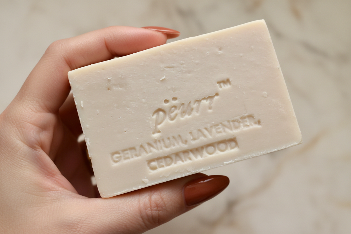 PËURR® Geranium, Lavender & Cedarwood soap held by a woman with long terracotta-copper nails against warm marble