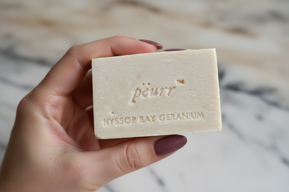 PËURR® Hyssop Bay Geranium soap held by a woman with a berry-plum manicure against warm marble