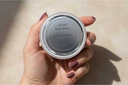 PËURR® Palmarosa Body Butter held by a woman with long berry-plum nails against warm marble