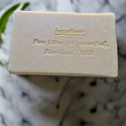 A rectangular bar of pale beige soap with embossed text reading 'Ingredients: Pure Olive Oil (saponified), Raw Goat's Milk', photographed close-up on a marble surface with a green leaf in the corner.