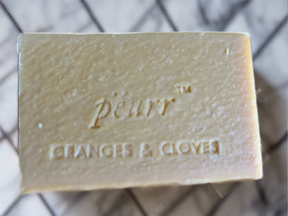 A bar of soap with 'peurr. ORANGES & CLOVES' engraved on it, with a light beige color and a textured surface.