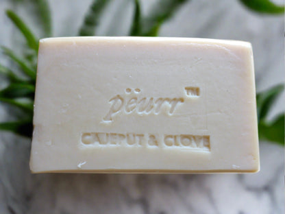 A bar of soap with 'PëURR CAJEPUT & CLOVE' embossed on it, placed on a white surface with green leaves in the background.