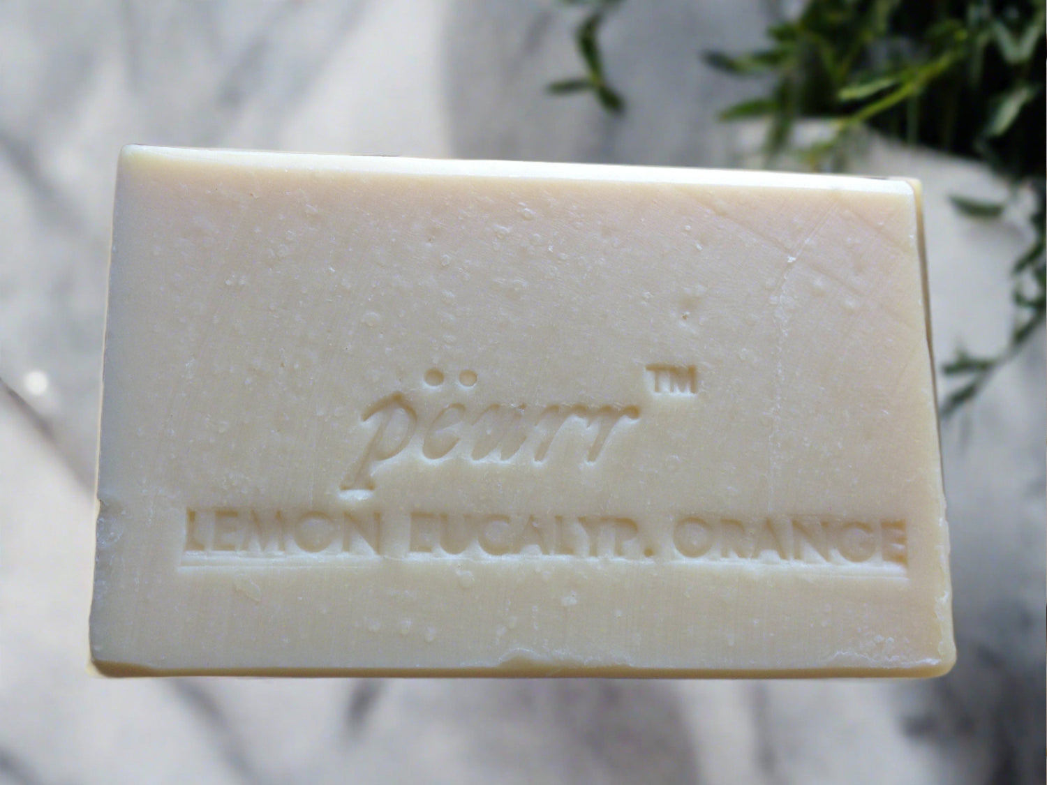 A bar of soap with 'PëURR LEMON EUCALYPTUS ORANGE' embossed on it, placed on a white surface with a eucalyptus leaf in the background.