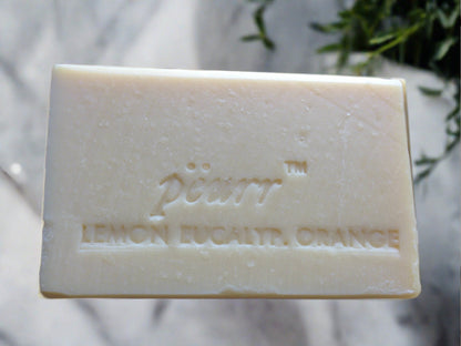 A bar of soap with 'PëURR LEMON EUCALYPTUS ORANGE' embossed on it, placed on a white surface with a eucalyptus leaf in the background.