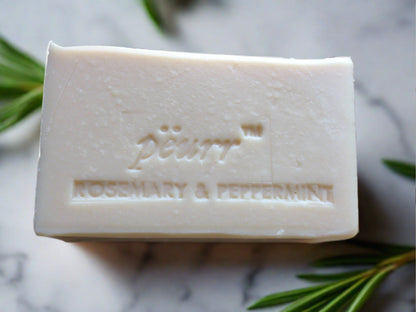 A bar of soap with 'Pëurr Rosemary & Peppermint' engraved on it, placed on a marble surface with a sprig of rosemary beside it.