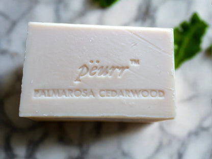 A bar of PÈURR® soap with 'PALMAROSA CEDARWOOD' engraved on it, placed on a marble surface with a few leaves of mint to the side.
