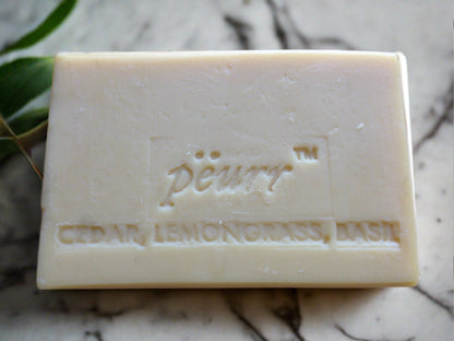 A bar of soap with 'Pëurr Cedar, Lemongrass, Basil' embossed on it, placed on a marble surface with green leaves in the background.

