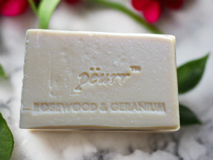 A bar of soap with 'PÉURR Rosewood & Geranium' embossed on it, placed on a surface with red flowers and green leaves in the background.