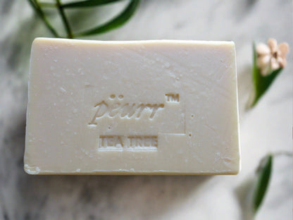 A bar of soap with 'PëURR® TEA TREE' embossed on it, placed on a surface with a few leaves and a flower around it.