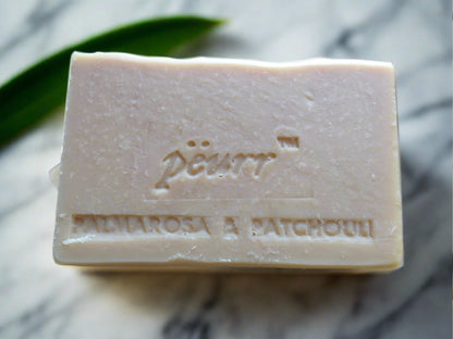 A bar of soap with 'PëURR' and 'PALMAROSA & PATCHOULI' embossed on the top, placed on a marble surface with a green leaf in the background.