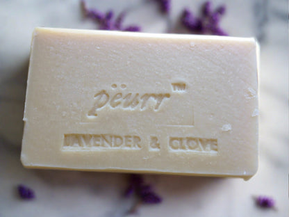 A bar of PÉURR branded soap with 'LAVENDER & CLOVE' engraved on it, surrounded by scattered lavender flowers.