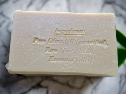 A beige bar of soap with the ingredients engraved on it with a marble background and some green leaves.