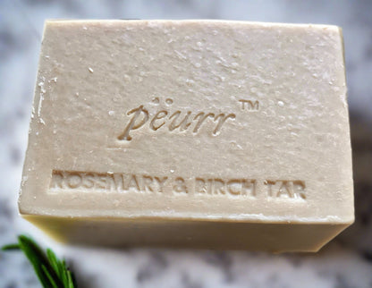 A bar of soap with 'Pëurr Rosemary & Birch Tar' engraved on it, presented alongside a sprig of rosemary.