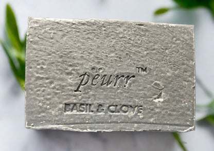 A bar of soap with 'peur' branding and 'basil & clove' text on it, set against a backdrop of green leaves.