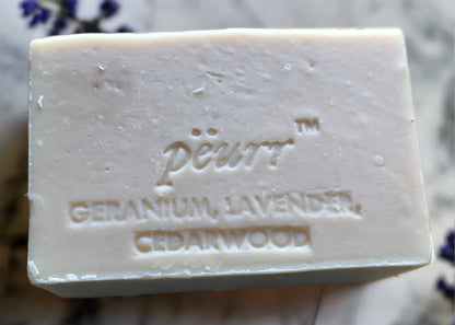A bar of soap with 'Pëurr GERANIUM, LAVENDER, CEDARWOOD' stamped on the bottom. The background has a floral pattern.