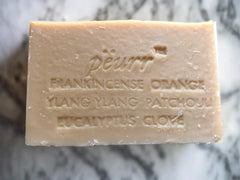 A bar of soap with the text 'PÈURR FRANKINCENSE ORANGE YLANG YLANG PATCHOULI EUCALYPTUS CLOVE' engraved on it.

