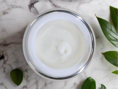 Jar of body butter with lid off showing creamy lotion flanked with green leaves.