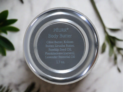 Bottom of body butter jar showing label with ingredients against light surface with green leaves