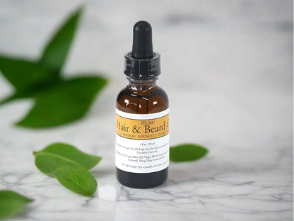 A bottle of PËURR Hair & Beard Oil with an orange label on a marble surface surrounded by green leaves.