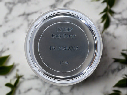 A jar of PëURR® Palmarosa Body Butter with the brand name and product title visible on the lid, placed on a surface with a leafy background.